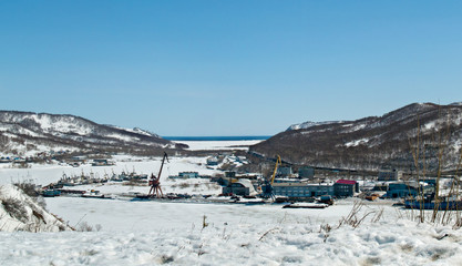 Fishing vessels and industrial buildings in winter in sunny weather near a frozen bay in Kamchatka