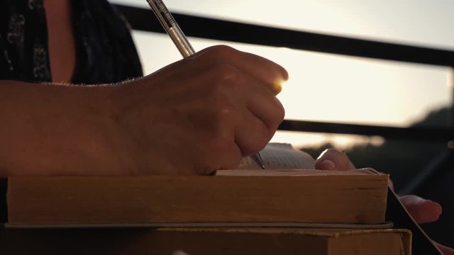 Woman's hand close-up, writing a note or letter to a notebook with a pen on a sunny day at sunset. A stack of scientific books, preparation for exams. Filling out the report
