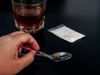 The hand of a person taking drug from a spoon with a syringe and a glass of whiskey Concept of drugs