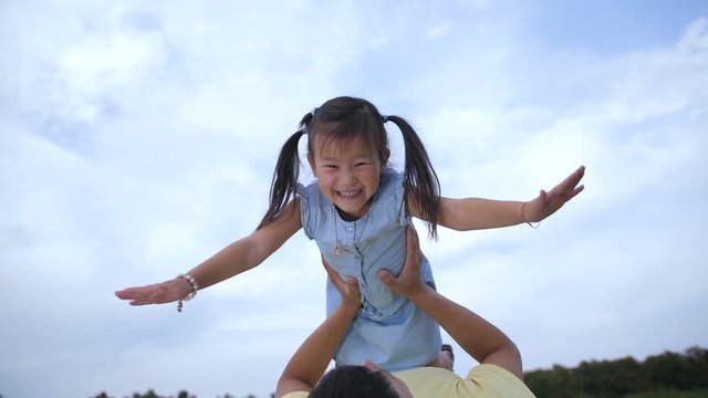 Portrait of carefree overjoyed little asian girl with pigtails making plane and laughing while loving father lifting up and keeping her above in summer park against blue sky background. Low angle view