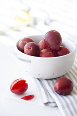 Red plums in white colander on a striped linen cloth with one plum sliced open in foreground