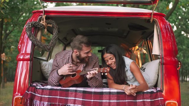 Happy young cheerful couple playing instruments and having some fun in retro hippie minibus in forest