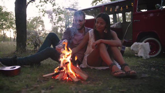 Beautiful young couple roasting marshmallows over a campfire while enjoying their road travel, slow motion, dolly