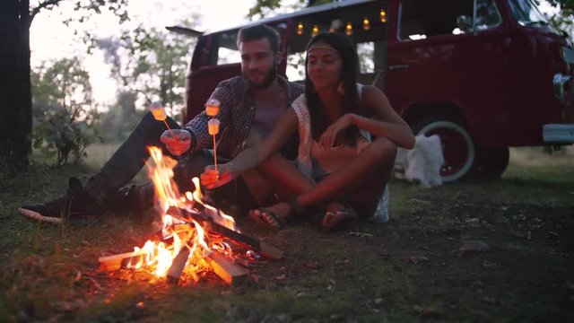 Beautiful young couple roasting marshmallows over a campfire while enjoying their road travel, slow motion, dolly