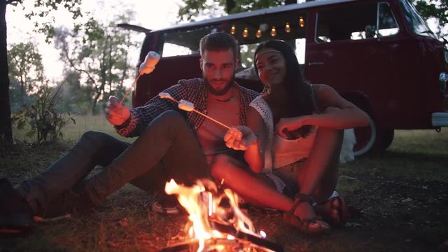 Beautiful young couple roasting marshmallows over a campfire while enjoying their road travel, slow motion