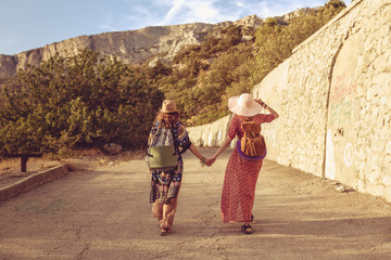 Fototapeta premium Girlfriends-travelers in the style of a jeeps are holding hands, brightly dressed with bonnies on their heads, walking along the road. In the background, mountains and forest. The concept of freedom.