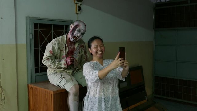 Asian woman taking photo with zombie actor by mobilr phone at haunted house in amusement park