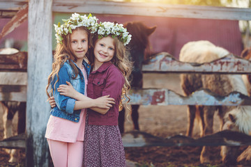 Two cute little girls with wreath of flowers playing on a haystack in countryside over sunset