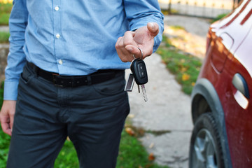 Man gives car keys outdoors with red car on background.