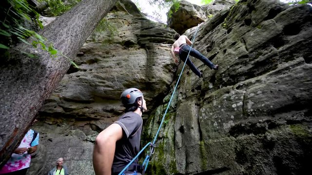 Young shirtless man rappels down a high rock after climbing to the top while his friend belays him.