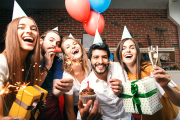 Group of friends holding gifts, sparklers and raising glasses with champagne, multicultural bearded man holding birthday cake, they are celebrating birthday