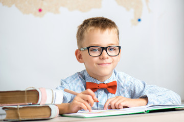 A schoolboy wears glasses, sits at a table, does homework and smiles.