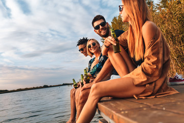 Group of young friends enjoying a day at the lake. They sitting on pier talking and drinking beers.