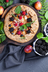 Whole-grain galette with plums and berries on dark background, top view
