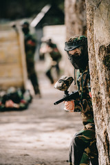 selective focus of female paintball player in goggle mask and camouflage aiming by paintball gun near tree outdoors