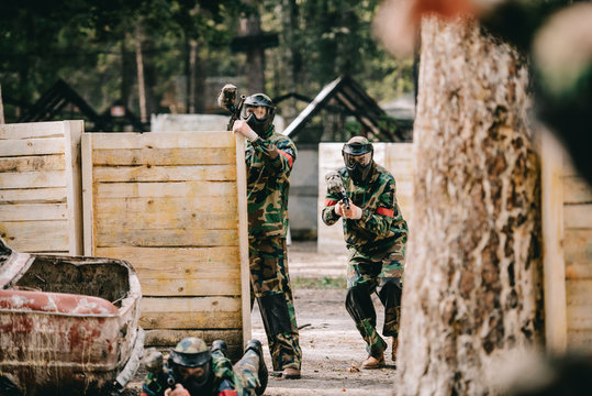selective focus of paintball team in uniform and protective masks playing paintball with marker guns outdoors