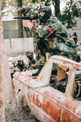 Fototapeta premium male paintball player aiming by marker gun and sitting on broken car while his team standing behind outdoors