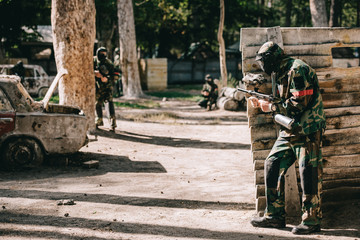 male paintball player hiding behind wooden wall while other team in uniform and protective masks standing with marker guns outdoors