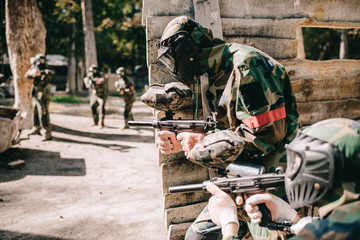 selective focus of paintball player in protective mask holding marker gun and his teammate hiding behind wooden wall outdoors