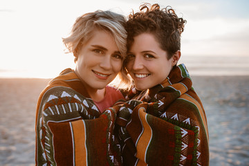 Smiling lesbian couple wrapped in a beach blanket at dusk