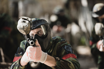 selective focus of serious male paintball player in goggle mask and camouflage aiming by paintball gun outdoors