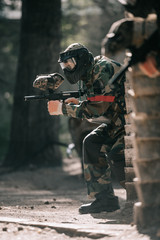 selective focus of male paintball player in goggle mask and camouflage aiming by paintball gun outdoors