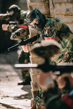 selective focus of female paintball player in camouflage and protective mask holding marker gun outdoors