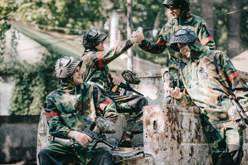 happy paintball team in camouflage celebrating victory and shaking hands on broken car outdoors