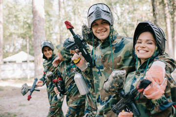 smiling young male paintballer embracing female teammate in camouflage with paintball gun outdoors - obrazy, fototapety, plakaty