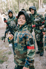 Fototapeta premium attractive smiling female paintballer in uniform holding paintball gun while her team standing behind outdoors