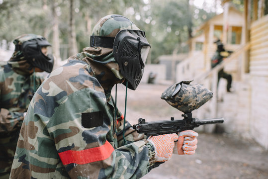 side view of male paintballer and his team in uniform and protective masks aiming by paintball guns outdoors
