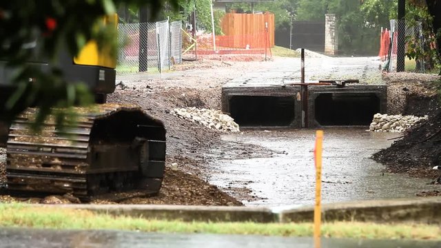 Heavy rain falls and collects in a ditch as a backhoe sits stationary with water droplets falling off it's tracks at a construction site.  Waterdrops bounce off the surface of the collecting water.