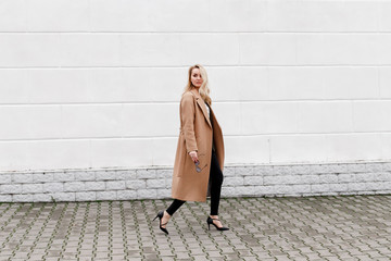 Beautiful young stylish blonde woman wearing long beige coat and black high heel shoes walking...