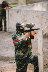 side view of confident male paintball player in goggle mask and camouflage aiming by paintball gun...
