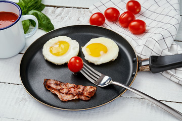 two delicious fried eggs and fried bacon in a pan on a white kitchen table with tomatoes juice and basil leaves