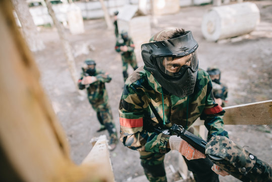 male paintball player in goggle mask and camouflage with marker gun on staircase while his team standing behind outdoors