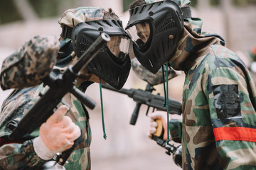 side view of male paintballers in protective masks and camouflage standing face to face outdoors