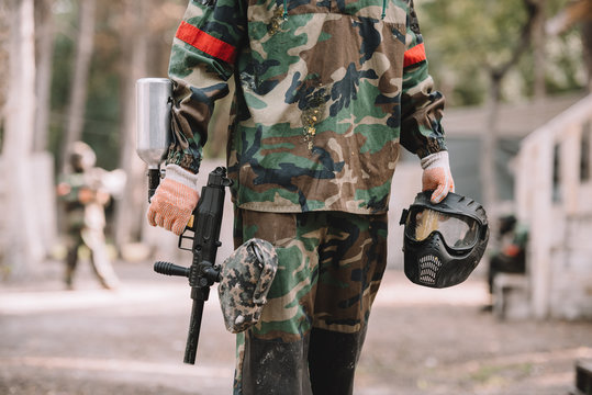 cropped image of paintball player holding marker gun and goggle mask covered by paintball splash outdoors