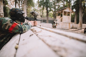 selective focus of paintball player in protective mask and camouflage aiming by marker gun outdoors