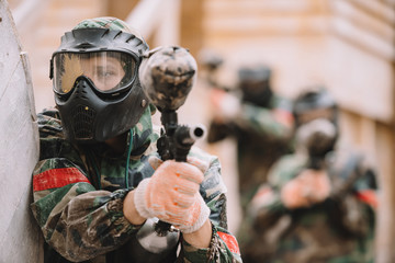 male paintballer in goggle mask covered by paintball splash aiming by marker gun outdoors