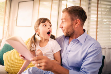 Dad is telling a story to his daughter in the living room at home and happy together. Setup studio shooting.