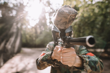 selective focus of male paintball player in goggle mask and camouflage aiming by paintball gun outdoors