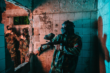 male paintball player in goggle mask and camouflage uniform hiding behind wall while other team is standing near in abandoned building