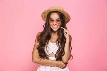 Photo of happy woman 20s wearing sunglasses and straw hat smiling at camera, isolated over pink background