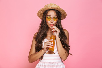 Photo of glamour woman 20s wearing sunglasses and straw hat drinking juice from glass bottle, isolated over pink background