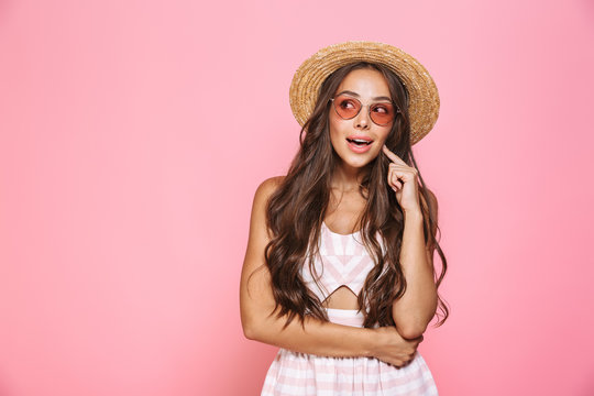 Photo of stylish woman 20s wearing sunglasses and straw hat smiling at camera, isolated over pink background