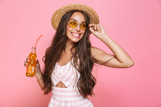 Photo of glamour woman 20s wearing sunglasses and straw hat drinking lemonade from glass bottle, isolated over pink background