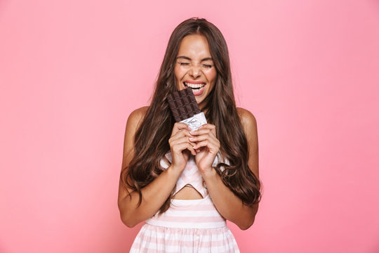Photo of joyous woman 20s wearing dress smiling and eating chocolate bar, isolated over pink background