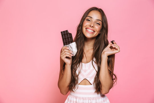 Photo of funny woman 20s wearing dress smiling and eating chocolate bar, isolated over pink background