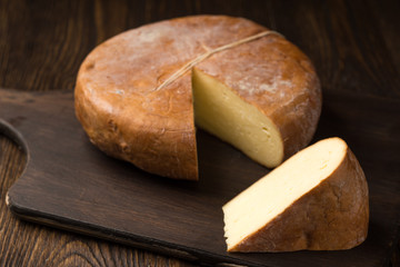 Cheese wheel and slice on a cutting board over a wooden table on natural environment.